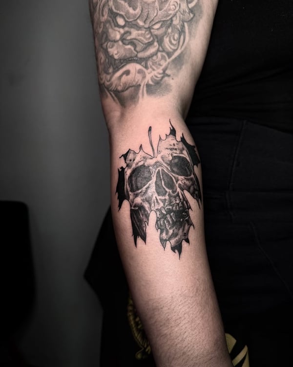 A black and grey skull tattoo with a leaf design on the forearm, featuring bold blackwork and illustrative style — by zealous, los angeles