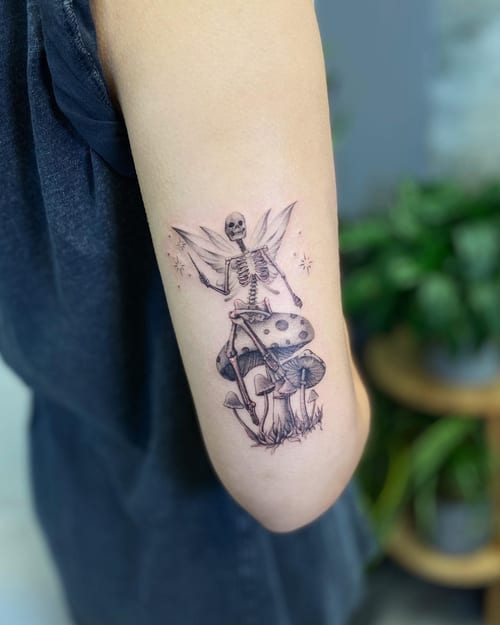 A whimsical black and grey tattoo of a skeleton with wings sitting on mushrooms, located on the arm — by Jian Liang, los angeles
