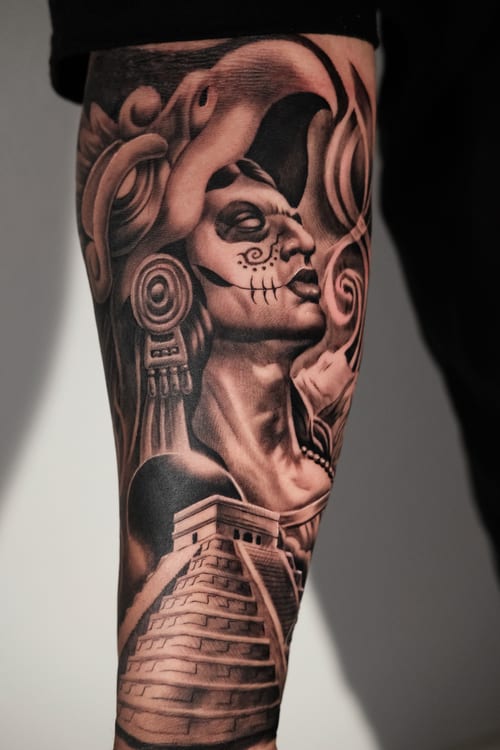 A detailed black and grey realism tattoo on the forearm depicting a mythological figure with intricate facial features and headdress, alongside an ancient pyramid — by Johnny Ayala, los angeles
