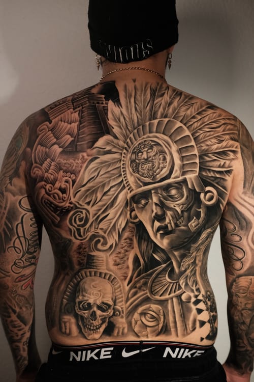 A detailed black and grey Chicano style tattoo covering the full back, featuring a mythological figure with a headdress and skull motifs — by Johnny Ayala, los angeles