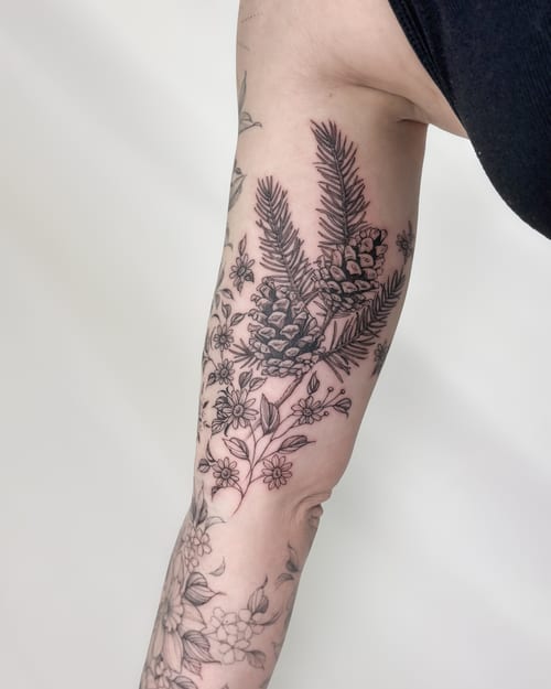 A delicate black and grey tattoo of pine cones and flowers on the inner arm, done in a fine line botanical style — by Melanie Benoit, los angeles