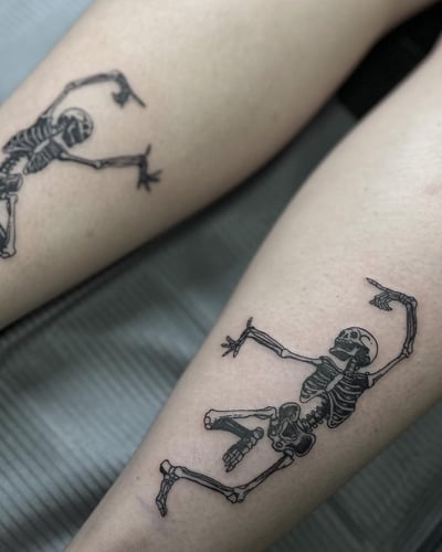 A fine line black and grey tattoo of dancing skeletons on the leg — by Maddie Ramirez, los angeles