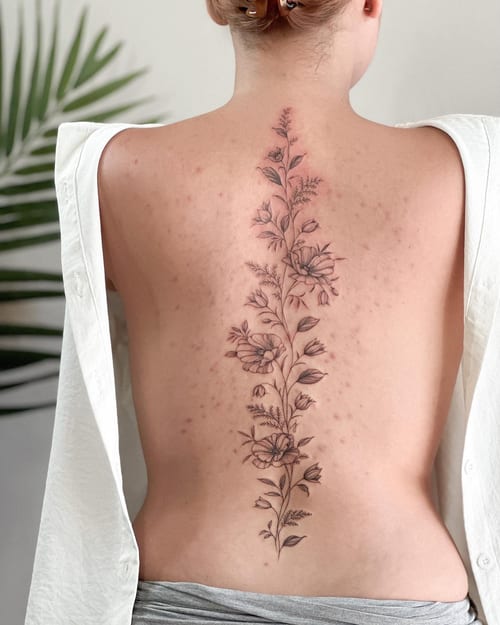 A delicate black and grey floral tattoo running vertically along the spine, featuring fine line work and minimalistic design — by Melanie Benoit, los angeles