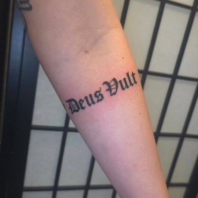 A bold black script lettering tattoo on the forearm reading 'Deus Vult' in a gothic style — by Miguel ANGELO, los angeles