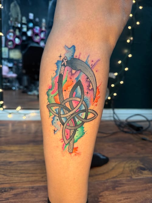 scythe woven through a celtic triquetra knot over rainbow watercolor splashes on the calf — by jessica linden, los angeles