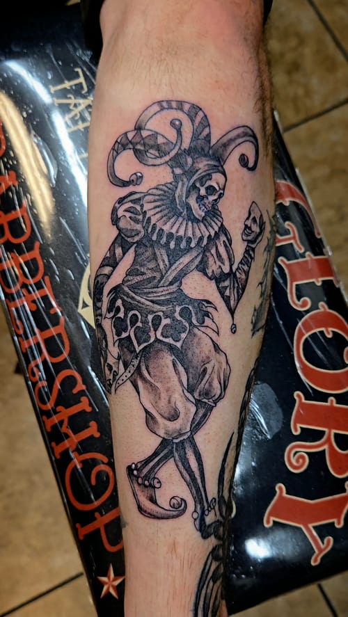 A black and grey illustrative tattoo of a jester on the forearm, featuring detailed linework and shading — by Dretty, los angeles