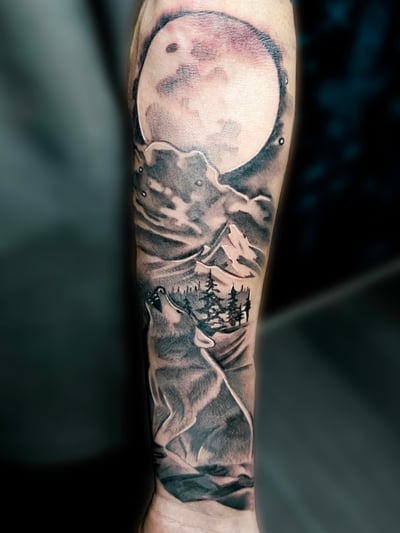 A black and grey realistic tattoo on the forearm depicting a howling wolf under a full moon with a mountainous landscape — by Josue Bravo, los angeles