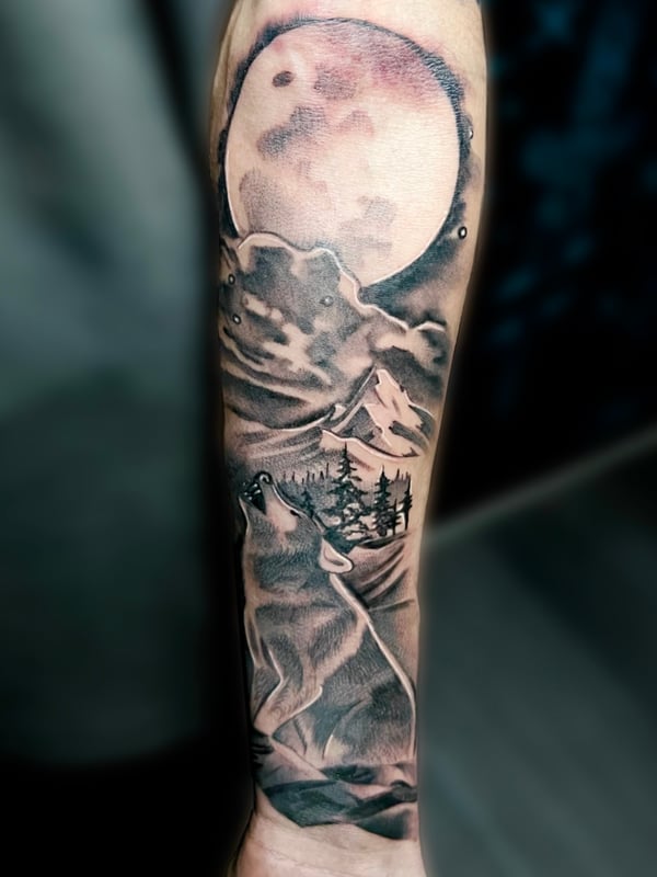 A black and grey realistic tattoo on the forearm depicting a howling wolf under a full moon with a mountainous landscape — by Josue Bravo, los angeles