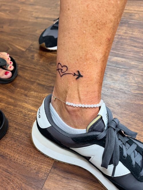 A minimalist black linework tattoo of a heart and airplane on the ankle, symbolizing travel and love — by Sandra Price, los angeles