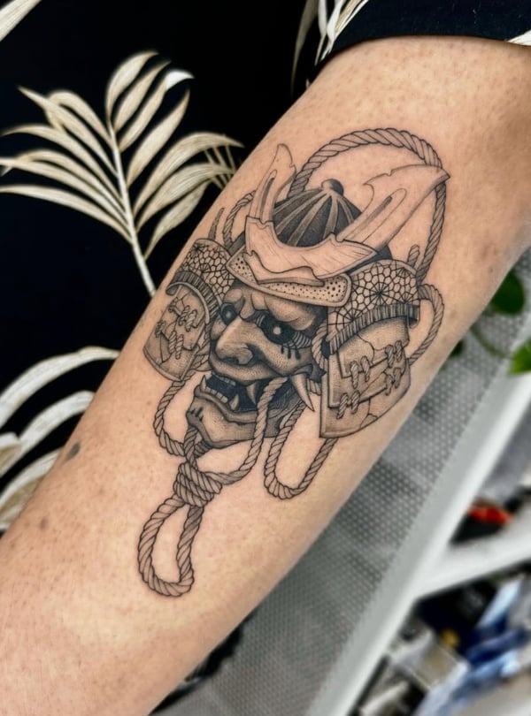 A black and grey Japanese style tattoo of a traditional samurai mask on the forearm, featuring intricate linework and shading — by otaybeatdown, orange, ca