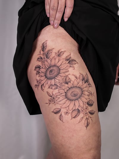 A delicate black and grey sunflower tattoo on the thigh with fine line detailing and floral elements — by Melanie Benoit, los angeles