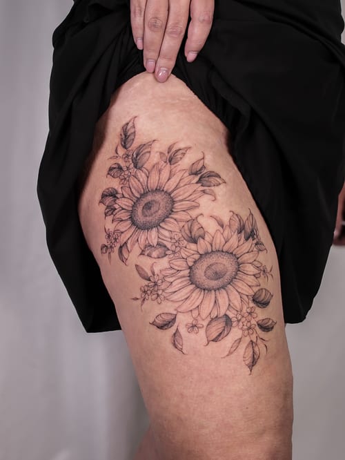 A delicate black and grey sunflower tattoo on the thigh with fine line detailing and floral elements — by Melanie Benoit, los angeles