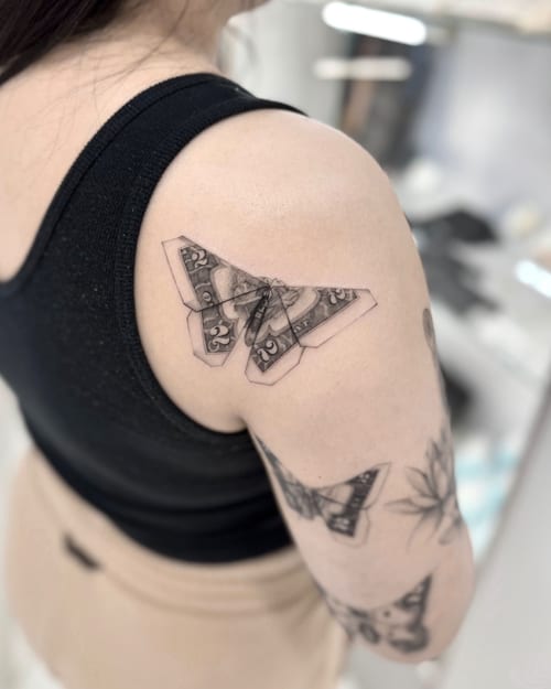 A black and grey minimalist tattoo of a butterfly made from folded currency on the shoulder, featuring clean lines and simple shading — by otaybeatdown, los angeles