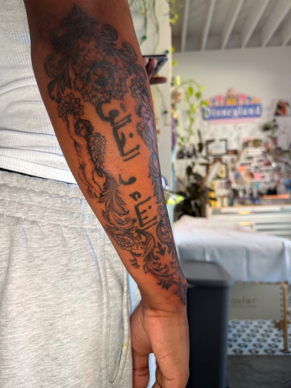 An ornate black and grey tattoo on the forearm featuring a cherub and script in an ornamental design — by Kiera Garvin, los angeles