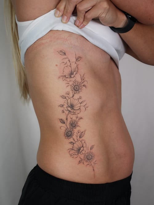 A delicate black and grey floral arrangement tattoo on the rib, featuring fine line work and minimalistic design — by Melanie Benoit, los angeles