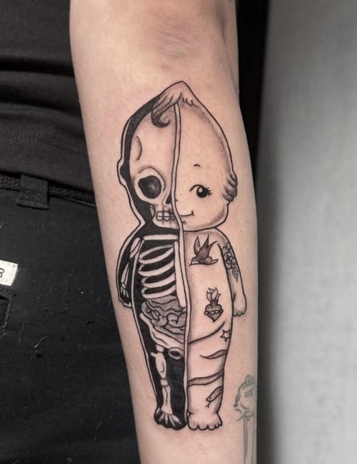 A black and grey illustrative tattoo on the forearm depicting a split figure with one half showing a skeleton and the other half a doll-like face — by Shelby Bianco, los angeles