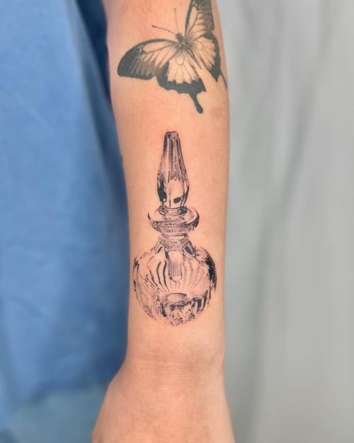 A black and grey fine line tattoo of a glass bottle on the forearm, with a butterfly above it, showcasing realistic detail and shading — by Level tattoo, los angeles