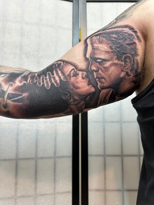 A black and grey realism tattoo of Frankenstein's Monster and the Bride of Frankenstein on the upper arm, showcasing detailed portraits and shading — by Humilde tattoos, los angeles
