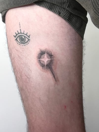 A minimalist black and grey tattoo featuring an eye and an abstract shape on the thigh — by Brian Kies, los angeles