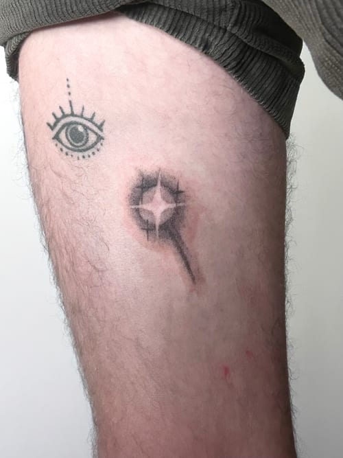A minimalist black and grey tattoo featuring an eye and an abstract shape on the thigh — by Brian Kies, los angeles