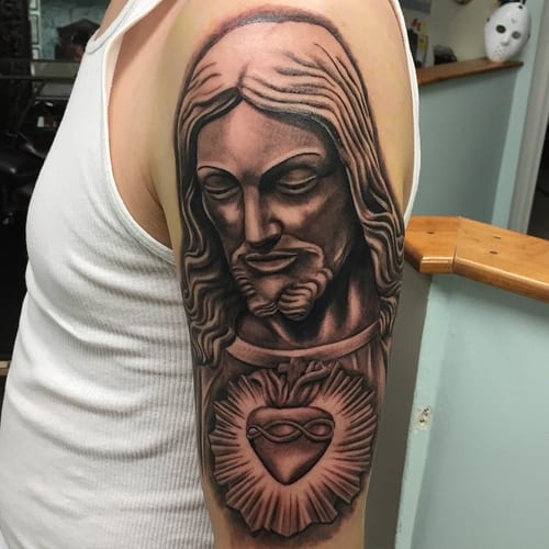 A realistic black and grey portrait of Jesus Christ with the Sacred Heart on the upper arm, featuring detailed shading and religious symbolism — by Johnny Ayala, los angeles