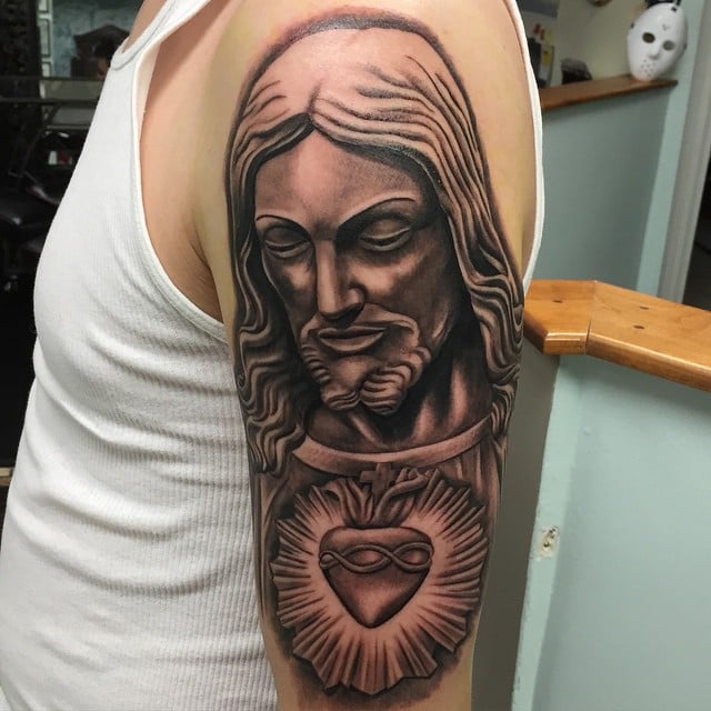 A realistic black and grey portrait of Jesus Christ with the Sacred Heart on the upper arm, featuring detailed shading and religious symbolism — by Johnny Ayala, los angeles