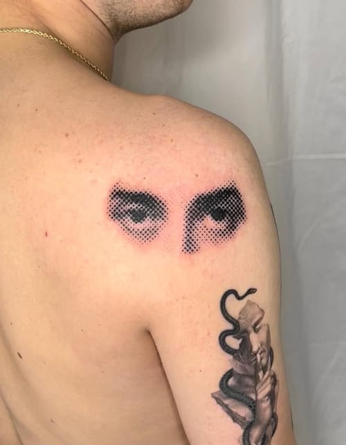 A black and grey dotwork tattoo on the shoulder featuring a pair of eyes and a snake wrapping around an arm. The eyes are rendered with a dotwork technique, creating a striking visual effect — by L…