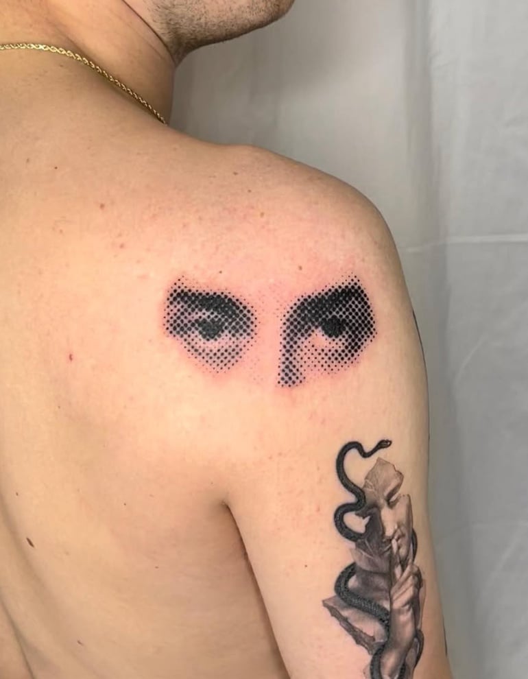 A black and grey dotwork tattoo on the shoulder featuring a pair of eyes and a snake wrapping around an arm. The eyes are rendered with a dotwork technique, creating a striking visual effect — by L…