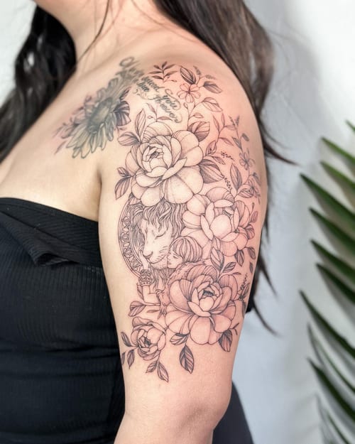 A delicate black and grey tattoo of a lion surrounded by flowers on the upper arm, featuring fine line details and ornate floral elements — by Melanie Benoit, los angeles
