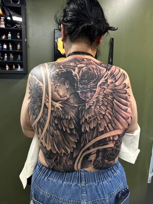 A detailed black and grey tattoo of an angel with wings and roses covering the full back — by Humilde tattoos, los angeles