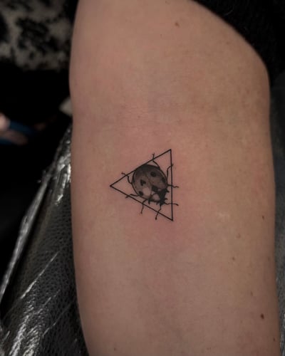 A small black and grey geometric tattoo on the arm featuring a moon within a triangle with barbed wire accents — by emily, los angeles