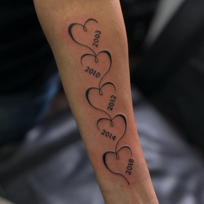 A minimalist blackwork tattoo of linked hearts with years on the forearm — by Maddie Ramirez, los angeles