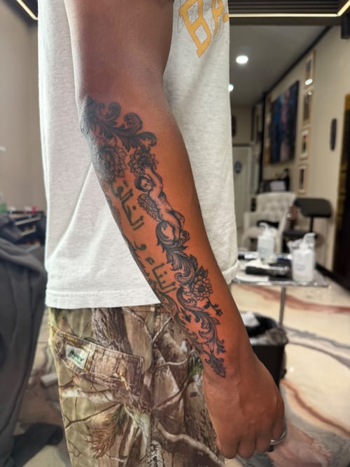 A black and grey cherub with floral design tattoo on the forearm, featuring ornate blackwork style — by Kiera Garvin, los angeles