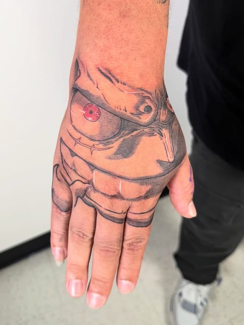 An anime character face tattoo on the hand with a mixed color profile, featuring bold black lines and red accents — by zealous, los angeles