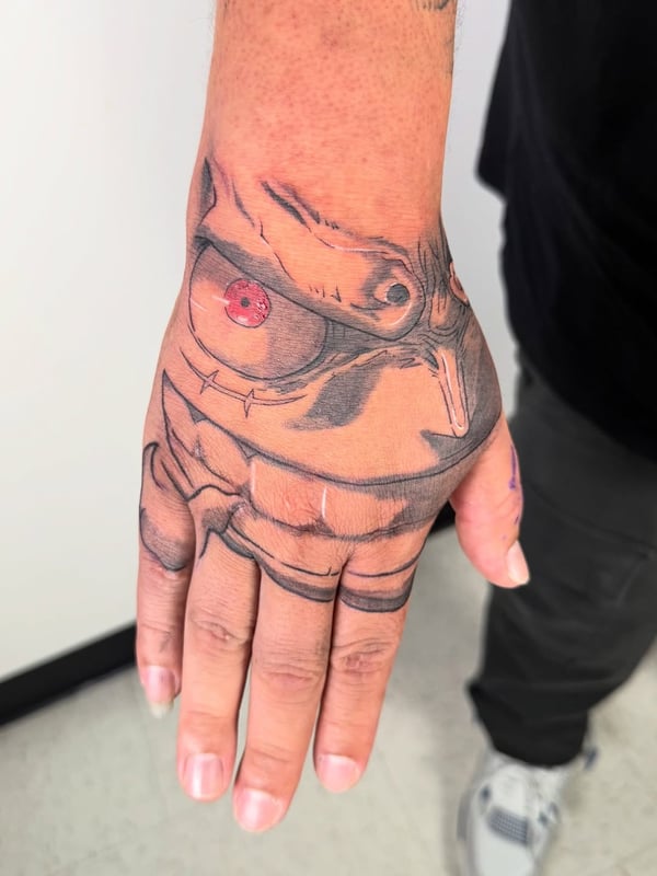 An anime character face tattoo on the hand with a mixed color profile, featuring bold black lines and red accents — by zealous, los angeles