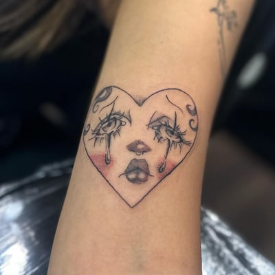 A delicate blackwork tattoo of a crying face in a heart shape on the arm — by Maddie Ramirez, los angeles