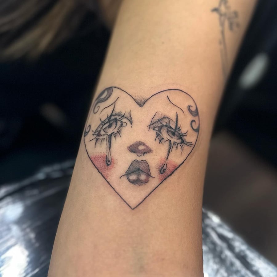 A delicate blackwork tattoo of a crying face in a heart shape on the arm — by Maddie Ramirez, los angeles