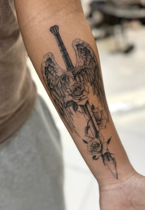 A black and grey tattoo on the forearm depicting a sword with angel wings and roses, rendered in a realistic style with detailed shading — by otaybeatdown, los angeles