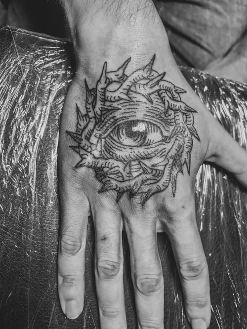A black and grey linework tattoo of an eye surrounded by thorn-like patterns on the hand, featuring bold outlines and intricate details — by Tag Devilish, los angeles