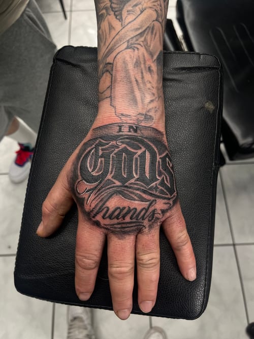 A black and grey lettering tattoo on the hand with bold script reading 'In God's Hands' — by Mr._Folks, los angeles
