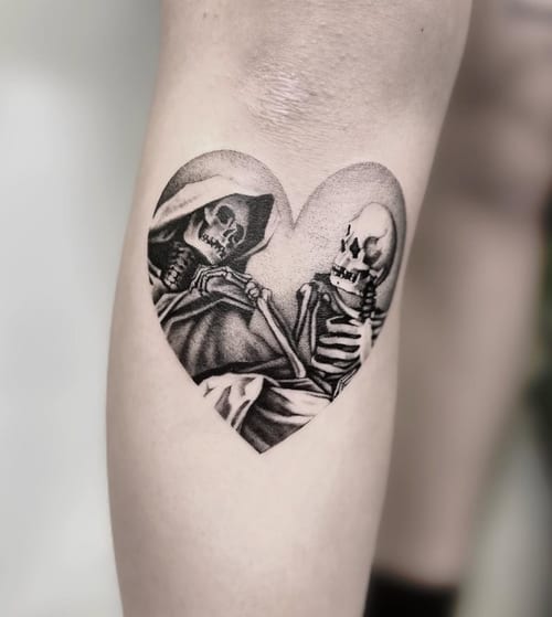 A black and grey tattoo of two skeletons inside a heart shape on the calf, featuring realistic shading and detail — by Jian Liang, los angeles