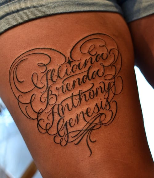 An elegant black script lettering tattoo on the thigh featuring multiple names in a delicate cursive style — by Johnny Ayala, los angeles
