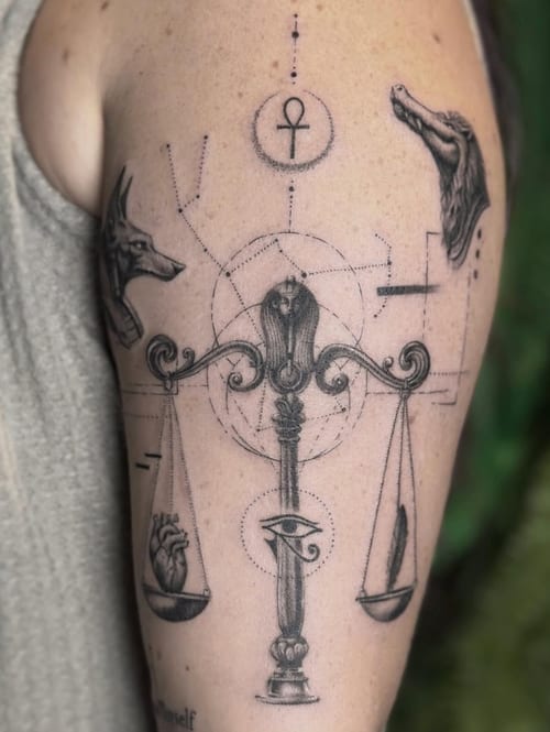 A black and grey tattoo on the upper arm featuring scales with a heart and feather, Anubis, and an Eye of Horus, using dotwork and linework styles — by Jian Liang, los angeles