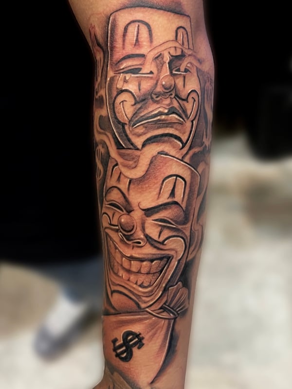 A black and grey Chicano style tattoo on the forearm featuring two theatrical masks with expressive faces and script elements — by Josue Bravo, los angeles