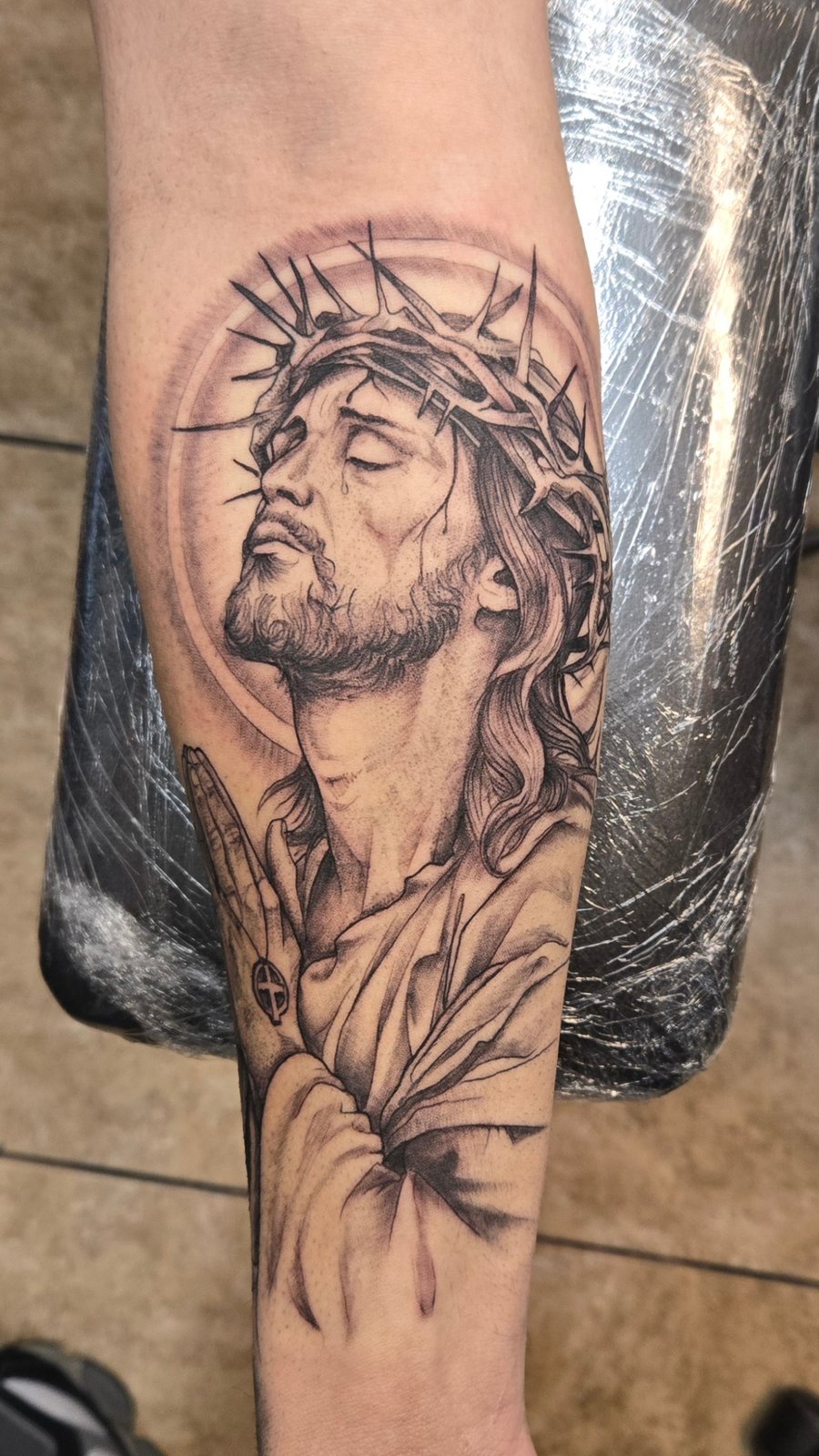 A detailed black and grey realism portrait of Jesus Christ with a crown of thorns on the forearm, featuring intricate shading and texture — by Dretty, los angeles