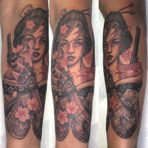 A detailed black and grey tattoo of a geisha holding a katana on the forearm, featuring intricate floral patterns and subtle color accents — by Sandra Price, los angeles