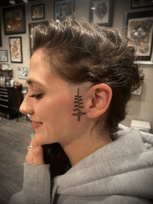 A minimalist black linework tattoo on the side of the face, featuring a simple geometric design — by tyler_whitlock, los angeles