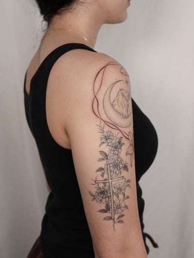 A delicate black and grey tattoo on the upper arm featuring a lion within a crescent moon and a floral cross design — by Melanie Benoit, los angeles