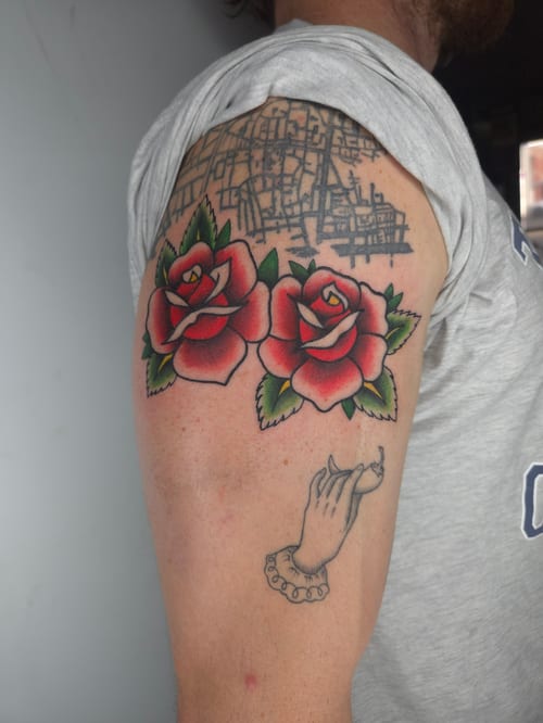 Two vibrant American Traditional style red roses on the upper arm, accompanied by an illustrative hand with a bracelet, all in bold colors — by ashynucklestattoo, los angeles