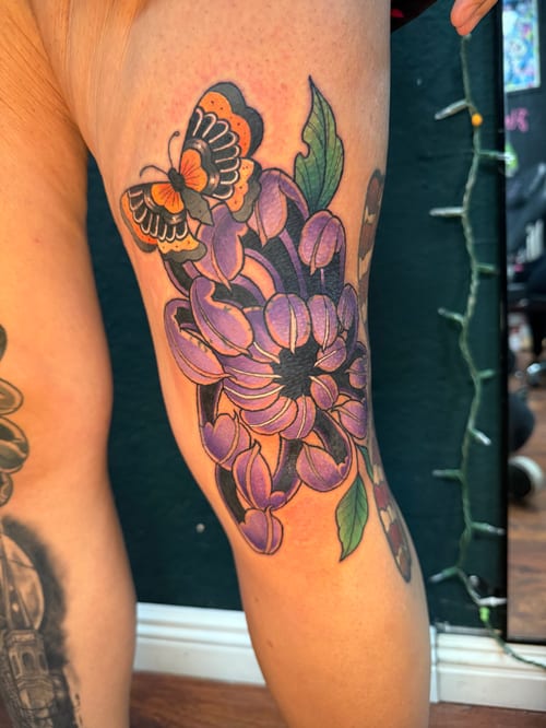 A colorful Neo-Traditional tattoo on the thigh featuring a large purple flower with green leaves and an orange butterfly, showcasing bold outlines and vibrant colors — by Jessica Linden, los angeles